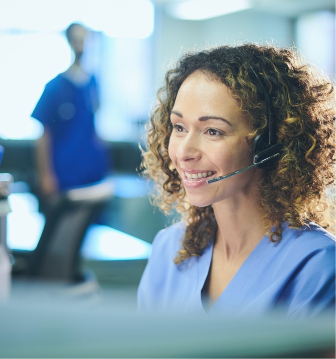 Nurse with curly hair delivering Patient Assistance Program support on a headset call, with another nurse working in the background.