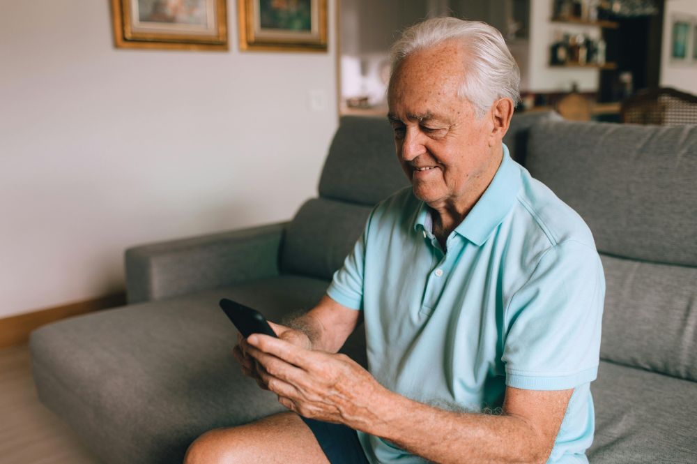 An older man sitting on a couch, using his phone for patient engagement in clinical trials.