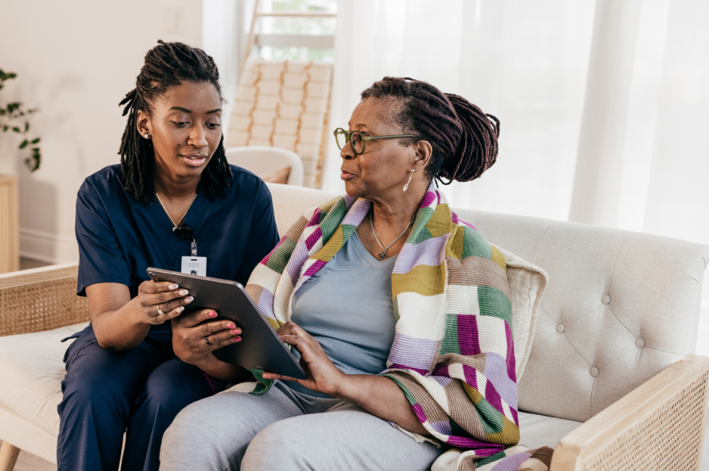 A nurse and an elderly woman sit on a couch, illustrating relationship-based patient recruitment in healthcare settings.